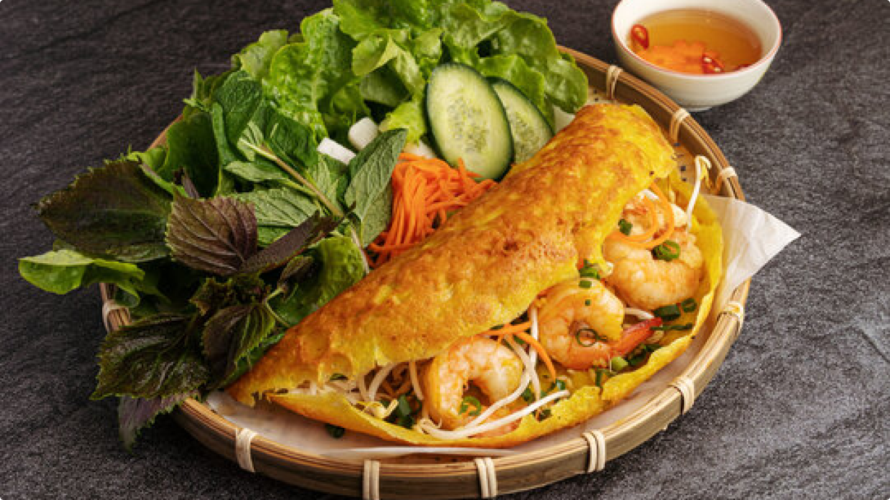 Image of a plate of banh xeo, filled with meat, vegetables and 3 shrimps. Sided with some vegetables. All placed on a nice bambo plate. Beside the place is a fish facuse dip