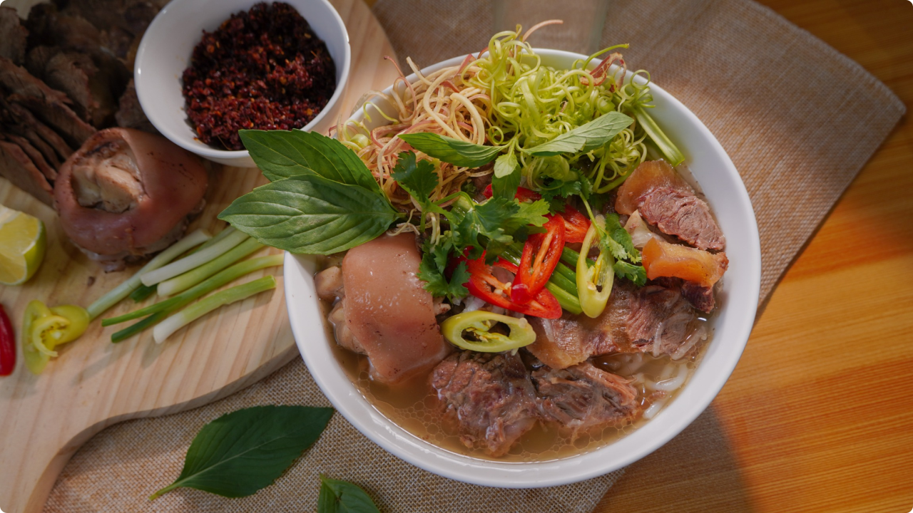 A bowl of Bún bò Huế with vegetables and meat on the bowl. Well placed on a table with side ekstra pig’s trotters and oxtail